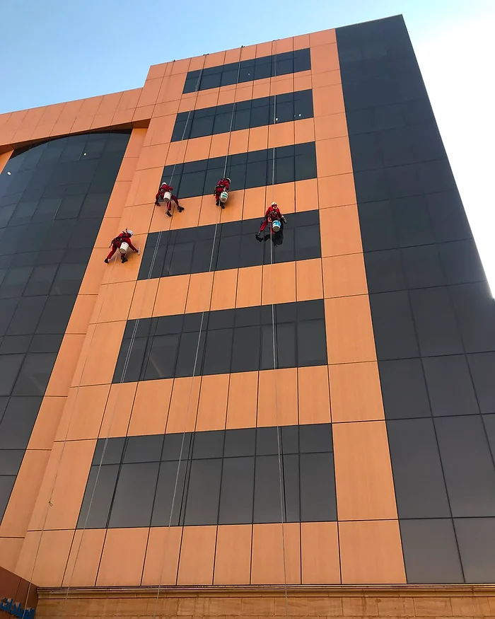 أربعة فنيين على حبال أمام واجهة من ألواح كلادينج برتقالية ورمادية